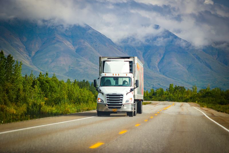 A white semi truck hauling cargo on a long stretch of road with mountains in the background